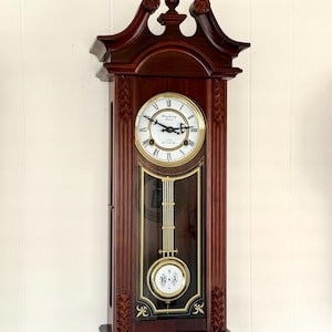 May include: A dark wood wall clock with a decorative pediment and finial. The white clock face features black Roman numerals and the text "Housbury Manor" and "8 Day Clock". A gold pendulum with the letters "R A" hangs behind a glass panel.