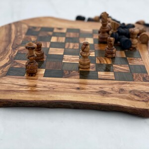May include: A wooden chessboard with a natural wood finish and a checkered pattern. The board is made of olive wood and has a unique, irregular shape. White and black chess pieces are scattered on the board.