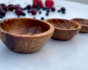 May include: Four small, round, wooden bowls made of olive wood. The bowls are arranged in a row, with the largest bowl in the front and the smallest bowl in the back. The bowls are all empty and have a natural wood finish.