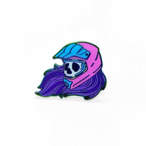May include: A pink and blue enamel pin featuring a skull wearing a motocross helmet with purple hair flowing behind it.