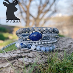 May include: A multi-strand bracelet with a large lapis lazuli gemstone, gray and blue stones, and leather cord. The bracelet is on a wooden log with green grass in the background.