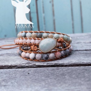Puede incluir: Un brazalete de envoltura multi-cadena con un colgante de piedra preciosa verde claro. El brazalete está hecho de cordón de cuero marrón y presenta cuentas de varios colores, incluyendo rosa claro, gris y blanco.