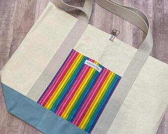 May include: Cream-colored tote bag with a light blue bottom and a rainbow-striped pocket. The bag has gray straps and a small tag that reads "FunkyCraftyThings". The bag is made of canvas-like material.
