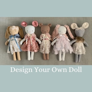 May include: Five handmade fabric dolls with different animal features, including a bunny, a mouse, a bear, and two other animals. The dolls are wearing lace dresses and have embroidered details. The dolls are arranged in a row on a light brown fabric background. The text "Design Your Own Doll" is displayed below the dolls.