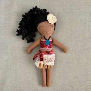 May include: A handmade doll with brown skin, black curly hair, and a white flower in her hair. She is wearing a red and white floral top, a white lace skirt, and a blue necklace.