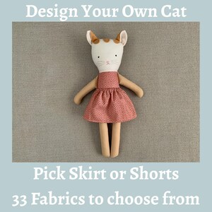 May include: A plush cat doll with a white face, tan ears, and embroidered features, wearing a dusty rose dress with a small white floral pattern. The text reads: Design Your Own Cat, Pick Skirt or Shorts, 33 Fabrics to choose from.