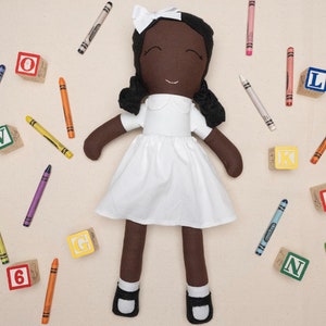 May include: A handmade doll with brown skin, black hair, and a white dress. The doll is surrounded by colorful crayons and wooden blocks with letters on them.
