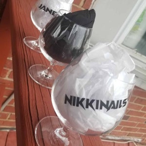 Puede incluir: Tres copas de tallo de vidrio transparente con letras negras. La primera copa dice "JANE". La segunda copa tiene un diseño negro. La tercera copa dice "NIKKINAILS".
