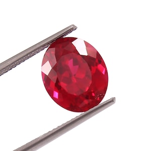 Puede incluir: Una gema ovalada de color rojo vibrante con un corte facetado, sostenida por unas pinzas.