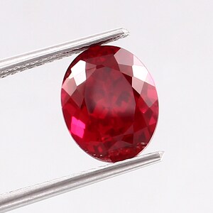 May include: A deep red oval-shaped gemstone with a faceted cut. The gemstone is held by a pair of tweezers.