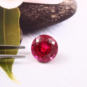 May include: A vibrant, deep red oval-shaped gemstone with a faceted cut. The gemstone is set against a white background.