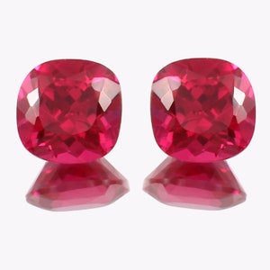 May include: Two vibrant red cushion-cut gemstones with a faceted finish. The gemstones are displayed on a white background with a reflection of the stones below.