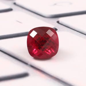 May include: A cushion-cut, deep red gemstone with a faceted surface that reflects light. The stone has a square shape with rounded corners and a vibrant, saturated color, displaying clear geometric patterns across its polished, multi-faceted top.