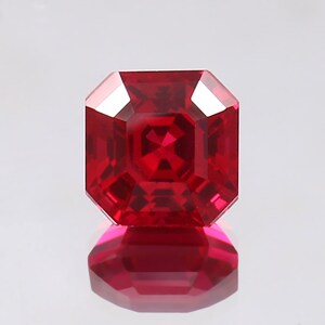 May include: A deep red gemstone with a square cut and a faceted finish. The gemstone is reflecting light, creating a bright shine.
