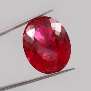 Puede incluir: Una gema ovalada de color rojo vibrante con un corte facetado. La gema está sujeta por unas pinzas sobre un fondo gris.