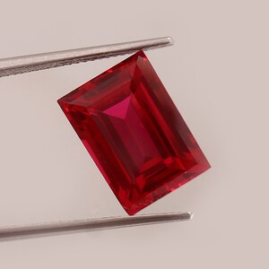 May include: A vibrant, rectangular-cut red gemstone with sharp, clean facets that reflect light. The stone displays a deep, saturated crimson hue throughout its structure, showcasing a clear, transparent quality and a polished, smooth surface finish.