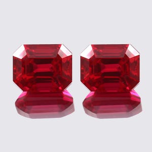 May include: Two vibrant red emerald-cut gemstones, each with a polished, faceted surface. The stones are displayed on a white background.