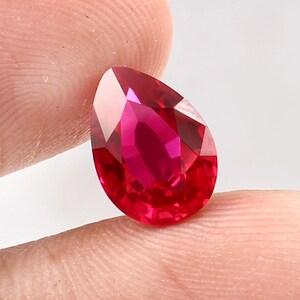 Könnte beinhalten: Ein birnenförmiger, facettierter Edelstein in einer leuchtenden, tiefroten Farbe. Der Stein hat eine polierte Oberfläche mit mehreren geometrischen Facetten, die das Licht reflektieren und für ein brillantes Aussehen sorgen. Der Edelstein wird zwischen zwei Fingern gehalten.