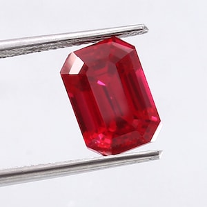 Puede incluir: Una piedra preciosa de color rojo oscuro tallada en esmeralda, sostenida por unas pinzas. La piedra preciosa es de un color rojo vibrante y tiene un alto nivel de claridad.