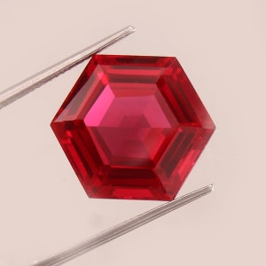 May include: A deep red hexagonal gemstone with a faceted cut. The gemstone is held by a pair of tweezers.