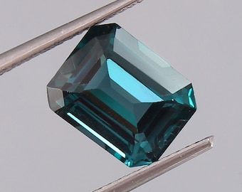 May include: A vibrant teal green gemstone with an emerald cut. The gemstone is held in a pair of tweezers.