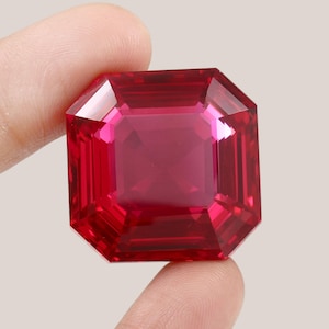 Puede incluir: Una gema roja vibrante con un corte cuadrado esmeralda. La gema está facetada y tiene un alto pulido.