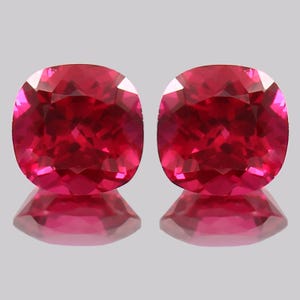 May include: Two cushion-cut, faceted gemstones with a deep red hue. The gems are displayed on a reflective surface, showcasing their brilliance and clarity. The shape is a square with rounded corners, and the color is a vibrant, saturated red.