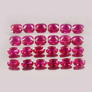 May include: A collection of 24 oval-shaped, faceted ruby gemstones. The rubies are a deep, rich red color and are arranged in three rows of eight.