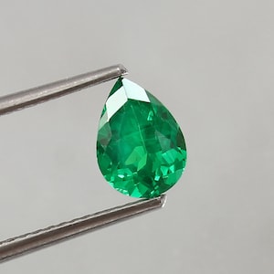 Op de afbeelding: Een peervormige smaragd edelsteen met een diepgroene kleur. De edelsteen wordt vastgehouden door een pincet tegen een witte achtergrond.