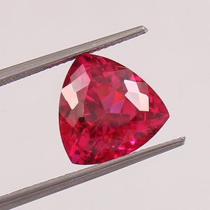 May include: A vibrant red triangular gemstone with a faceted cut, held by tweezers.