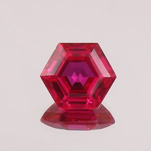 May include: A faceted, hexagonal gemstone with a deep red hue. The gem's facets reflect light, creating a brilliant display. The gem is set against a white background, with its reflection visible below.