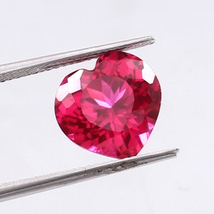 May include: A heart-shaped, faceted ruby gemstone with a deep red color. The gemstone is held in tweezers.