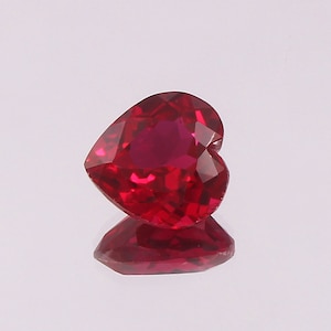 May include: A heart-shaped, faceted gemstone with a deep, vibrant red color. The stone features a polished surface with multiple geometric cuts that reflect light, creating a brilliant appearance. A clear reflection of the gem is visible below.