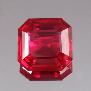 Könnte beinhalten: Ein facettierter, rechteckig geschliffener Rubin-Edelstein. Der Edelstein hat eine leuchtend rote Farbe mit scharfen, sauberen Kanten und einer reflektierenden Oberfläche. Der Edelstein wird auf einer reflektierenden Oberfläche präsentiert.