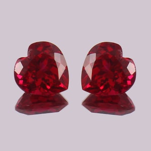 May include: Two heart-shaped ruby gemstones, each with a deep red colour and a brilliant shine. The gemstones are set against a light grey background.