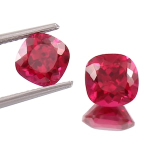 Op de afbeelding: Twee kussenvormig geslepen, gefacetteerde, rode edelstenen. Eén wordt vastgehouden door een zilveren pincet, de andere rust op een reflecterend oppervlak en toont zijn facetten. De edelstenen hebben een levendige, dieprode kleur en een vierkante vorm met afgeronde hoeken.