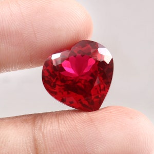 Peut inclure: Une pierre précieuse en forme de cœur, facettée, d'un rouge vif. La gemme est tenue entre deux doigts, mettant en valeur ses surfaces polies et les reflets de la lumière sur ses facettes. La couleur de la pierre est d'un rouge profond.
