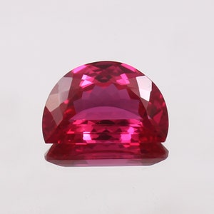 May include: A vibrant magenta gemstone, cut in a semi-circular shape. The facets of the gem catch the light, creating a sparkling effect. The polished surface reflects light, highlighting its brilliance. The gemstone is set against a plain white background.