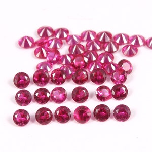 May include: A collection of round, faceted, ruby red gemstones. The gemstones are arranged in a loose pile on a white surface.