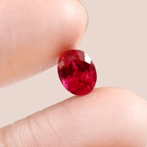 May include: A vibrant, oval-cut ruby gemstone with a deep red color and multiple facets that reflect light. The stone displays a clear, polished surface with internal depth and a brilliant, saturated crimson hue throughout its entire structure.