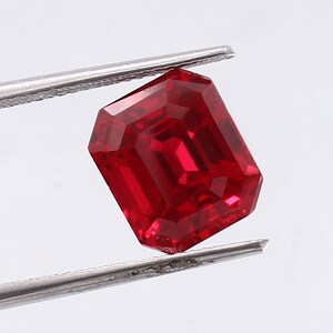 May include: A deep red emerald cut gemstone held by tweezers. The gemstone is a vibrant red color and has a high level of clarity.