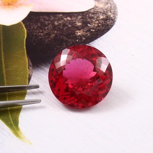 Pode incluir: Uma gema redonda e vermelha com corte facetado. A gema está colocada sobre um fundo branco.