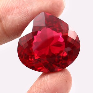 May include: A vibrant, deep red gemstone cut into a heart shape with numerous facets that reflect light. The stone displays a rich, saturated crimson hue and a high-clarity, transparent appearance, showcasing a precise geometric cut.