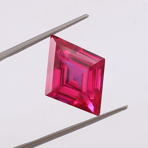 Può includere: Una gemma color rosa intenso a forma di aquilone con una superficie sfaccettata che crea riflessi di luce geometrici. La pietra è tenuta da una pinzetta in metallo argentato, che ne mette in risalto i bordi affilati e il colore.