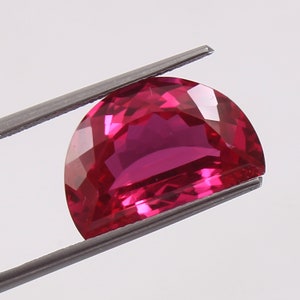 Op de afbeelding: Een gefacetteerde, halfronde, dieproze edelsteen, vastgehouden door een metalen pincet. De edelsteen heeft een gepolijst oppervlak en een briljante slijpvorm, die licht reflecteert. De achtergrond is effen wit.