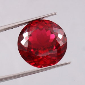 Puede incluir: Una piedra preciosa de color rojo intenso con forma redonda y superficie facetada. La piedra preciosa está sujeta por unas pinzas.