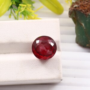 May include: A deep red gemstone with a faceted cut, displayed on a white surface.