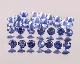 May include: A collection of loose blue sapphires, some are faceted and some are round, arranged in a pyramid shape on a white surface.