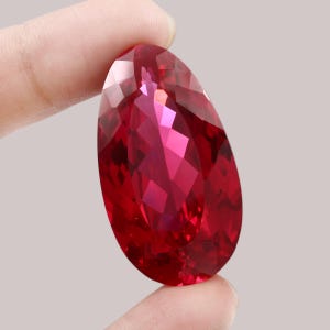 Peut inclure: Une pierre précieuse à facettes en forme de poire d'une couleur rouge profond et vibrante. La pierre présente de nombreuses facettes géométriques qui reflètent la lumière, créant un aspect brillant et étincelant sur toute sa surface.
