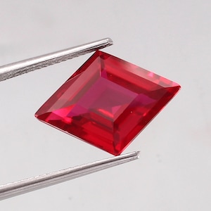 May include: A square-cut, deep red gemstone held by silver tweezers. The facets of the gem catch the light, highlighting its rich colour and clarity. The gem is a saturated, vibrant red.